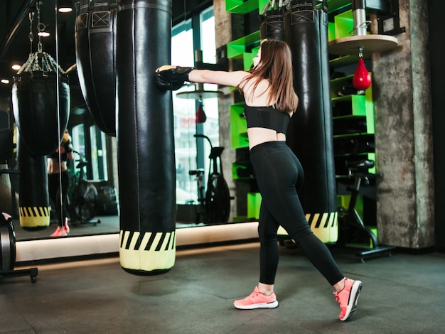 Encajar chica en ropa deportiva y guantes de boxeo entrena golpes contra un saco de boxeo en un gimnasio oscuro