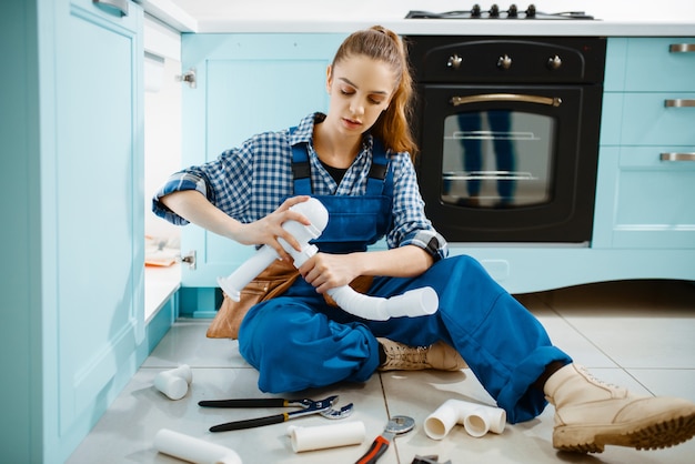 Encanador feminino atraente no problema de fixação uniforme com tubo de drenagem na cozinha. Handywoman com pia de conserto de bolsa de ferramentas, serviço de equipamento sanitário em casa