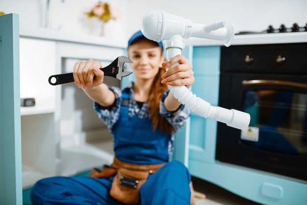 Encanador feminino jovem de uniforme mostra a chave inglesa e o tubo na cozinha. handywoman com pia de conserto de bolsa de ferramentas, serviço de equipamento sanitário em casa