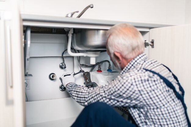 Encanador sênior trabalhando com ferramentas de encanamento na cozinha. Renovação.