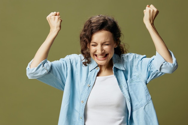 Encantada, emocionada, feliz, rizada, hermosa dama con pantalones vaqueros, camisa casual, levanta las manos con alegría, posando aislada en un fondo verde oliva, pastel, siendo uno mismo, gente, estilo de vida, emociones, concepto, espacio de copia.