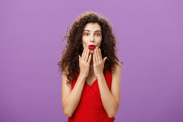 Encantada y emocionada mujer adulta de pelo rizado de lujo con maquillaje de noche en vestido rojo doblando los labios con asombro sosteniendo las palmas en la cara escuchando atentamente un rumor interesante sobre la pared púrpura.