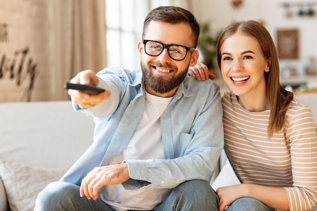 Encantado casal adulto homem e mulher sorrindo e olhando para a câmera enquanto está sentado no sofá e mudando de canal na TV com controle remoto