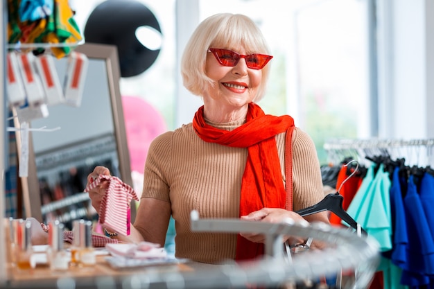 Encantado com a coleção da loja. Retrato de uma senhora idosa alegre olhando para longe com óculos de sol vermelhos enquanto escolhe uma bandana para combinar com seu vestido