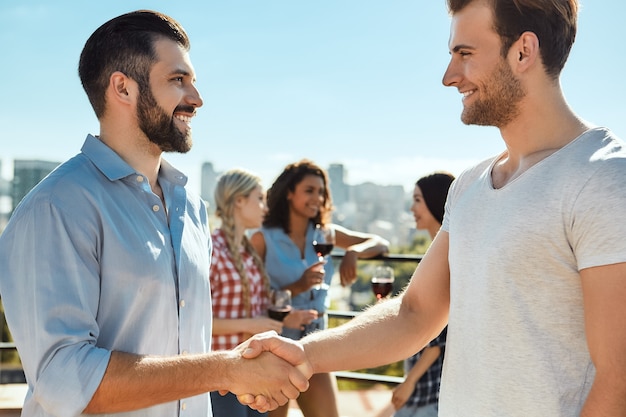 Foto encantado de conocerlos, dos jóvenes felices dándose la mano y sonriendo mientras están de pie en