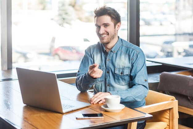 Encantado jovem sorridente satisfeito trabalhando no laptop apontando o dedo para a câmera