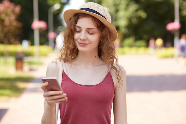 Encantado joven feliz con expresión facial agradable, sonriendo sinceramente, leyendo noticias, sosteniendo un teléfono inteligente