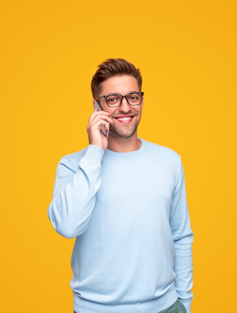 Encantado joven con gafas sosteniendo la mano en el bolsillo y mirando a la cámara con una sonrisa mientras tiene una conversación por teléfono inteligente contra el fondo amarillo