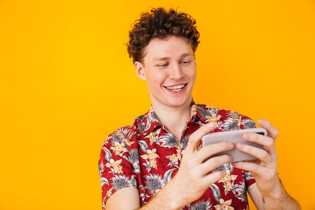 Encantado joven hombre jugando videojuegos en el teléfono móvil y sonriendo aislado en amarillo
