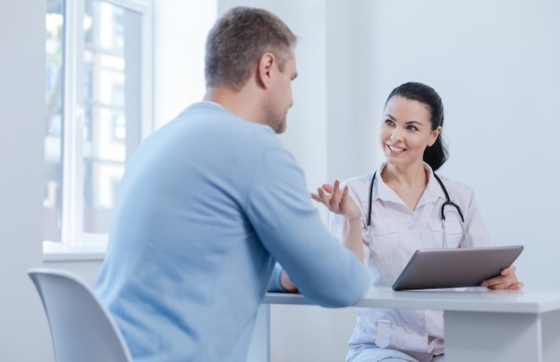 Encantado médico hábil involucrado disfrutando de una cita en el hospital y usando un dispositivo digital mientras expresa felicidad