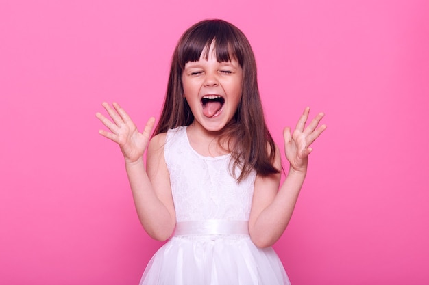 Encantado de niña linda con un vestido blanco gritando felizmente, gritando con los ojos cerrados y la boca abierta, asombrada, aislada sobre la pared rosa