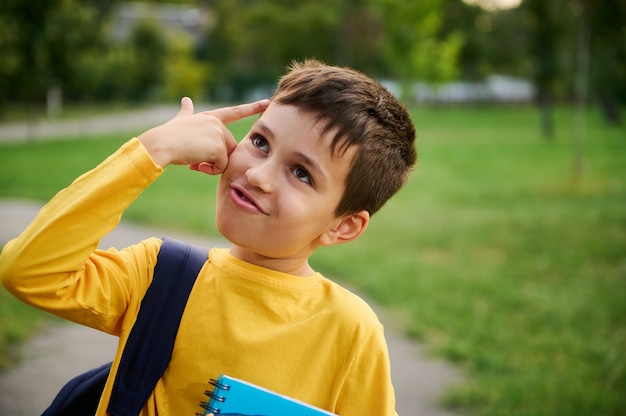 Un encantador y alegre colegial alegre se lleva la mano a la sien imitando una pistola, como un signo de cansancio por estudiar después de un duro día en la escuela, mirando hacia el fondo del parque de la ciudad