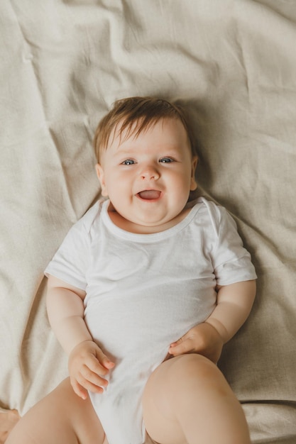 El encantador bebé de seis meses de ojos azules yace en la cama con un traje blanco.