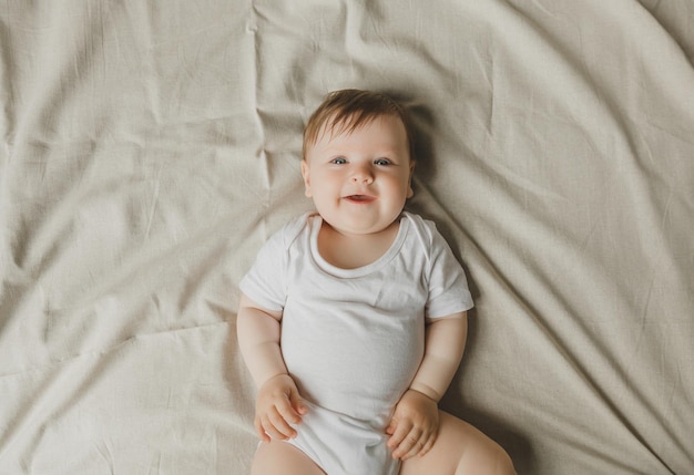 El encantador bebé de seis meses de ojos azules yace en la cama con un traje blanco.