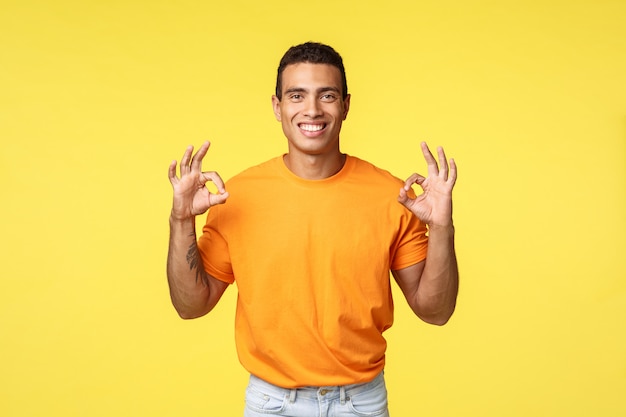 Encantador chico feliz con brazo tatuado en camiseta naranja, pantalón blanco, muestra bien