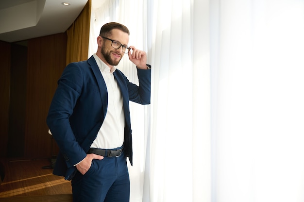 Encantador y exitoso hombre de negocios, próspero inversor de negocios, joven emprendedor durante un viaje de negocios, se para en la ventana de una habitación de hotel y sonríe mirando a la cámara a través de anteojos