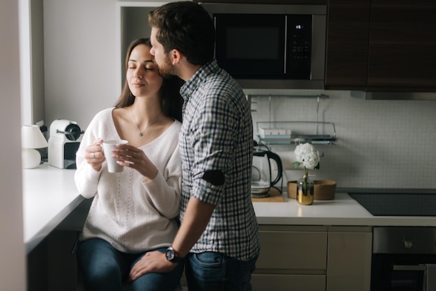 Encantador hombre feliz está besando a su novia en la cocina junto a la ventana. La vida diaria de un hombre y una mujer en el apartamento.