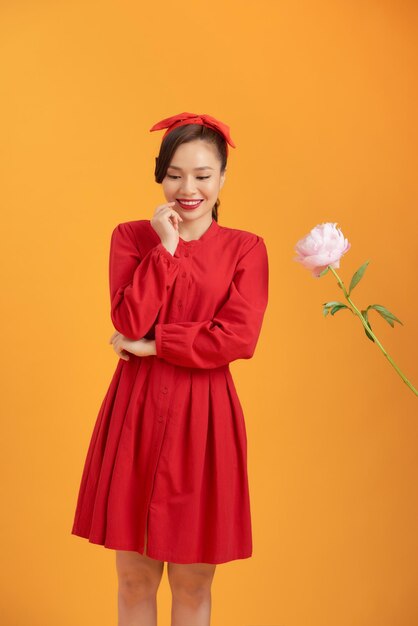 Encantador joven asiático vestido rojo sosteniendo una flor de peonía aislada sobre un fondo naranja