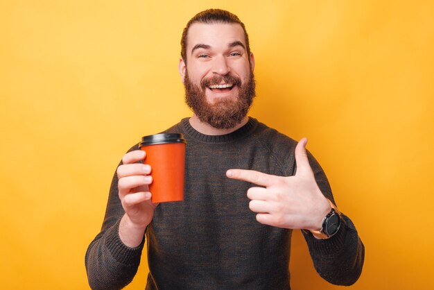 Encantador joven barbudo apuntando a la taza de café roja para llevar