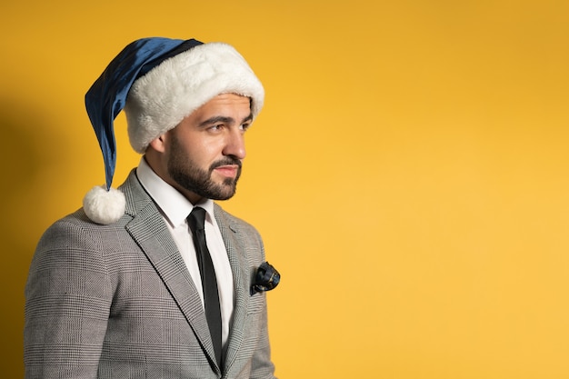 Foto encantador joven barbudo con sombrero azul de santa y traje gris, mirando hacia los lados aislado en la pared amarilla