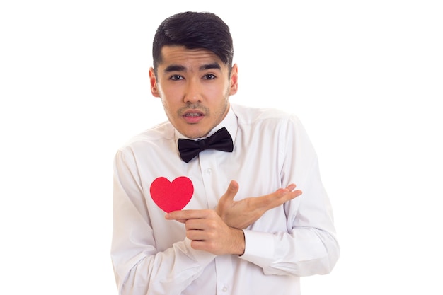 Encantador joven con cabello oscuro en camiseta blanca con corbatín negro sosteniendo un corazón de papel rojo