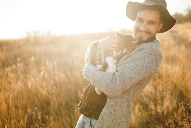 Encantador joven hipster con un gato Un chico con bigote y una hermosa sonrisa está abrazando a un gato