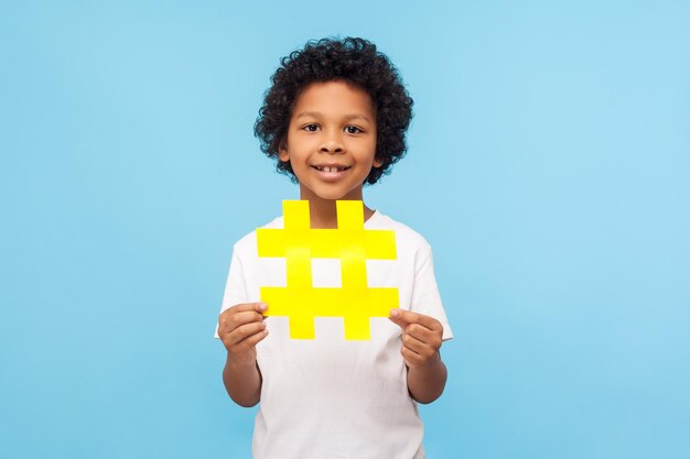 Encantador menino bonitinho feliz com cabelo encaracolado em t-shirt segurando o grande símbolo de hashtag amarelo e sorrindo para a câmera, blogueiro infantil mostrando sinal de hash. tiro de estúdio interno isolado em fundo azul