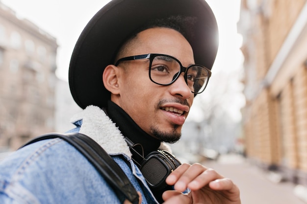 Encantador modelo masculino africano posando con expresión pensativa en el fondo de la ciudad. Foto de primer plano de joven negro lleva gafas y sombrero de moda.