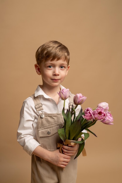 un encantador niño de cuatro años con overoles beige y una camisa blanca sostiene un ramo de tulipanes rosas