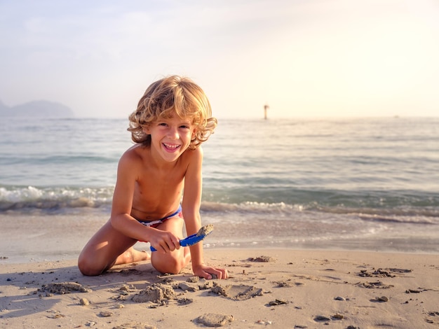 Encantador niño de pelo rizado jugando con una pala de plástico en una playa de arena húmeda bañada por un mar claro y mirando a la cámara con una sonrisa feliz