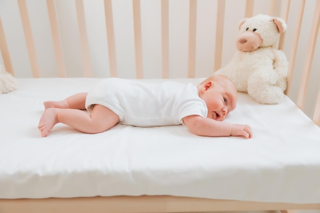 Un encantador y sonriente bebé de 2 meses con ojos azules y un traje blanco yace en una cuna junto a un oso de peluche Vista desde arriba de un recién nacido