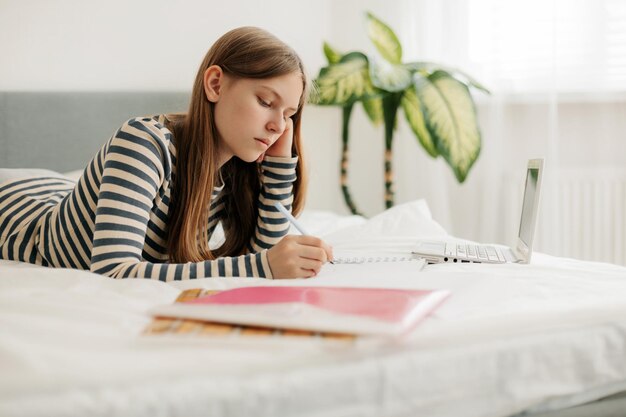 Foto una encantadora adolescente está acostada en la cama haciendo la tarea con una computadora portátil tomando notas