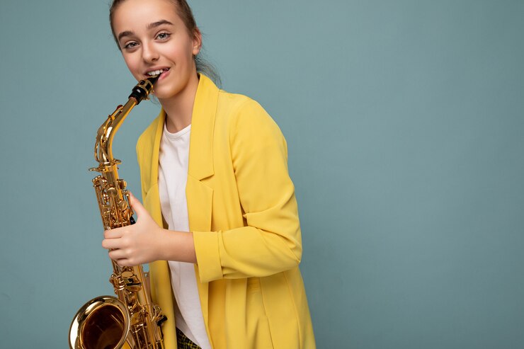Foto encantadora adolescente morena sonriente positiva con chaqueta amarilla de moda que se encuentran aisladas sobre la pared de fondo azul tocando el saxofón mirando a la cámara. copia espacio