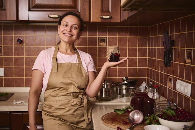 Encantadora y agradable pastelera sostiene un tarro de confitura de cereza casero en el acogedor interior de la cocina rústica