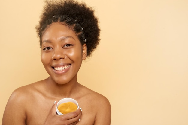 Encantadora y alegre mujer afroamericana rizada sostiene parches cosméticos debajo de sus ojos mira con alegría las poses de la cámara en un espacio de copia de fondo beige para su anuncio