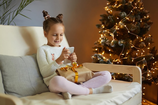 Una encantadora y alegre niñita con suéter sentada en una tos blanca cerca del árbol de Navidad en el interior de una casa sosteniendo una caja de regalo para Navidad y usando un teléfono celular haciendo videollamadas o viendo dibujos animados