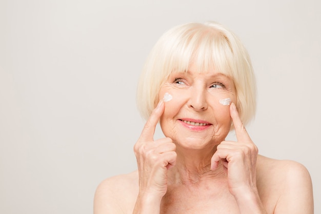 Foto encantadora, bonita, anciana tocando la piel suave perfecta de su rostro con los dedos, sonriendo a la cámara sobre fondo gris, usando crema facial de día y noche, procedimientos de cosmetología