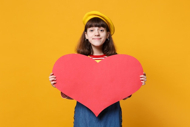 Encantadora chica adolescente con boina francesa, vestido de mezclilla sosteniendo un gran corazón rojo vacío aislado en el fondo de la pared amarilla en el estudio. Emociones sinceras de la gente, concepto de estilo de vida. Simulacros de espacio de copia.