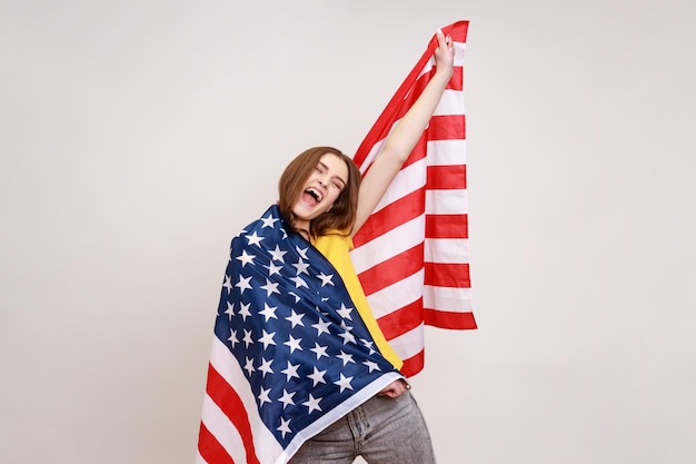 Encantadora chica adolescente bonita con cabello ondulado, envolviendo la bandera estadounidense, gritando alegremente, levantando la mano, celebrando el día de la Independencia. Disparo de estudio interior aislado sobre fondo gris.
