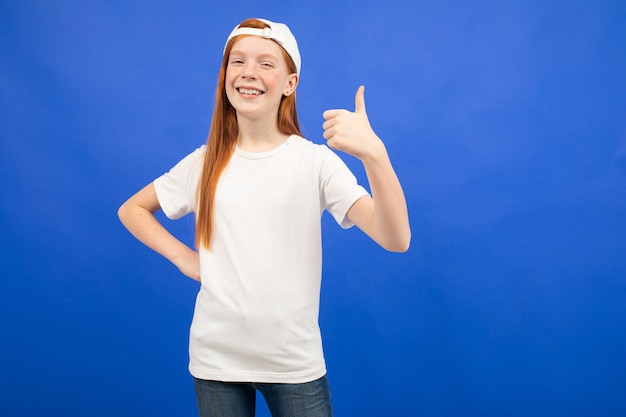 Encantadora chica adolescente pelirroja en una camiseta blanca con un diseño que muestra una super clase pulgares arriba azul