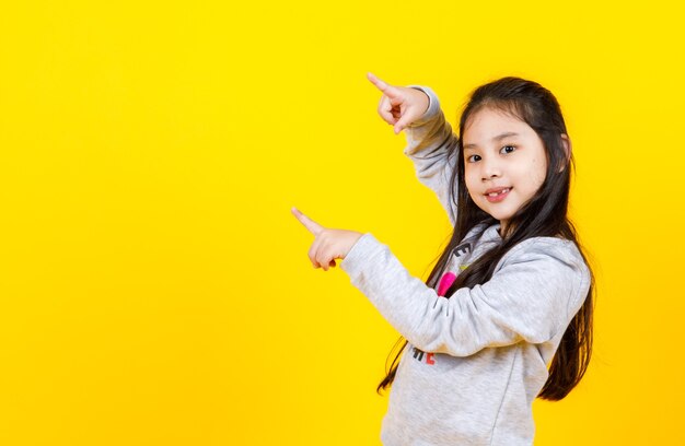 Encantadora chica asiática con suéter sonriendo y disfruta gesticulando con el signo de la mano para señalar con ambos dedos el mensaje deseado en el espacio de la copia para recomendar la oferta de beneficios de invierno del retrato aislado de un niño feliz