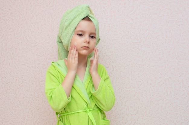 Encantadora chica en bata verde después de la ducha se aplica crema en la cara, en las mejillas. Concepto de cuidado de la piel.