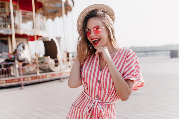 Encantadora chica blanca con sombrero de paja riéndose mientras posa al lado del carrusel Impresionante jovencita rubia pasando el día de verano en el parque de diversiones