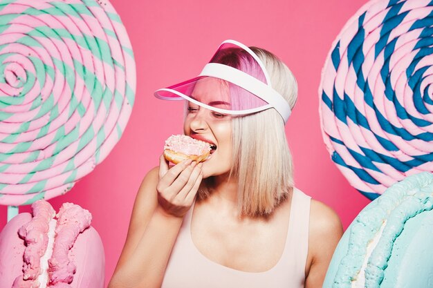 Encantadora chica con cabello rubio vistiendo top de pie con enormes paletas de dulce en estudio rosa