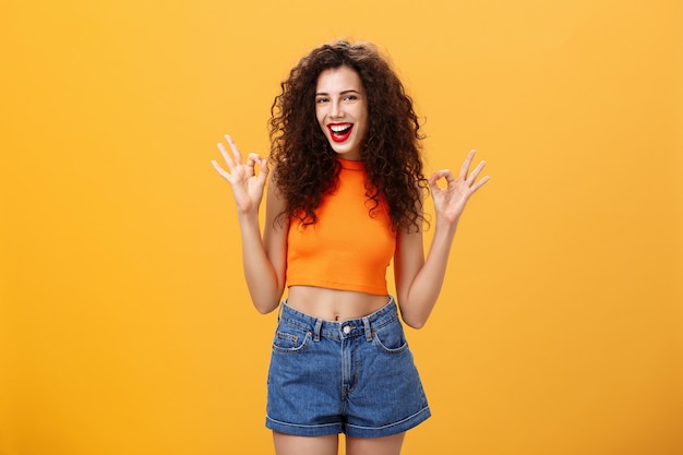 Encantadora chica caucásica extrovertida y amigable con peinado rizado en top recortado y pantalones cortos mostrando un gesto de aprobación expresando que le gusta una fiesta increíble sonriendo a la cámara sobre la pared naranja.