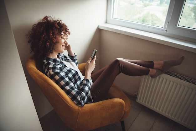 Foto encantadora chica caucásica con pelo rizado sentado en el sillón y sonríe mientras charla en el móvil