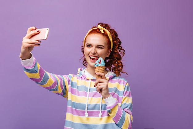 Encantadora chica con diadema amarilla guiña un ojo, sostiene helado y toma selfie en pared púrpura