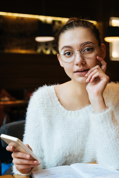 Encantadora chica hermosa con gafas y un suéter descansando en un café después del trabajo sosteniendo un teléfono inteligente