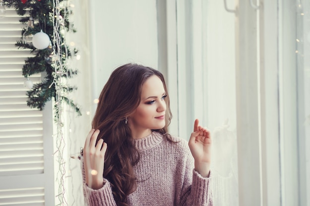 Encantadora chica hermosa en suéter sentado en la ventana de Navidad