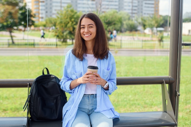 Encantadora chica hipster esperando el autobús o el tranvía en la estación de transporte público en la mañana con una taza de café